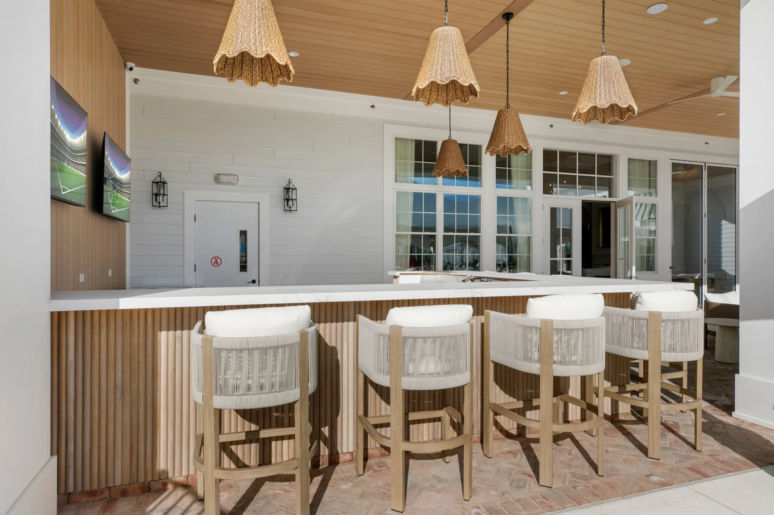 Covered outdoor bar with a fluted wood front, woven pendant lights, upholstered bar stools and wall-mounted TVs