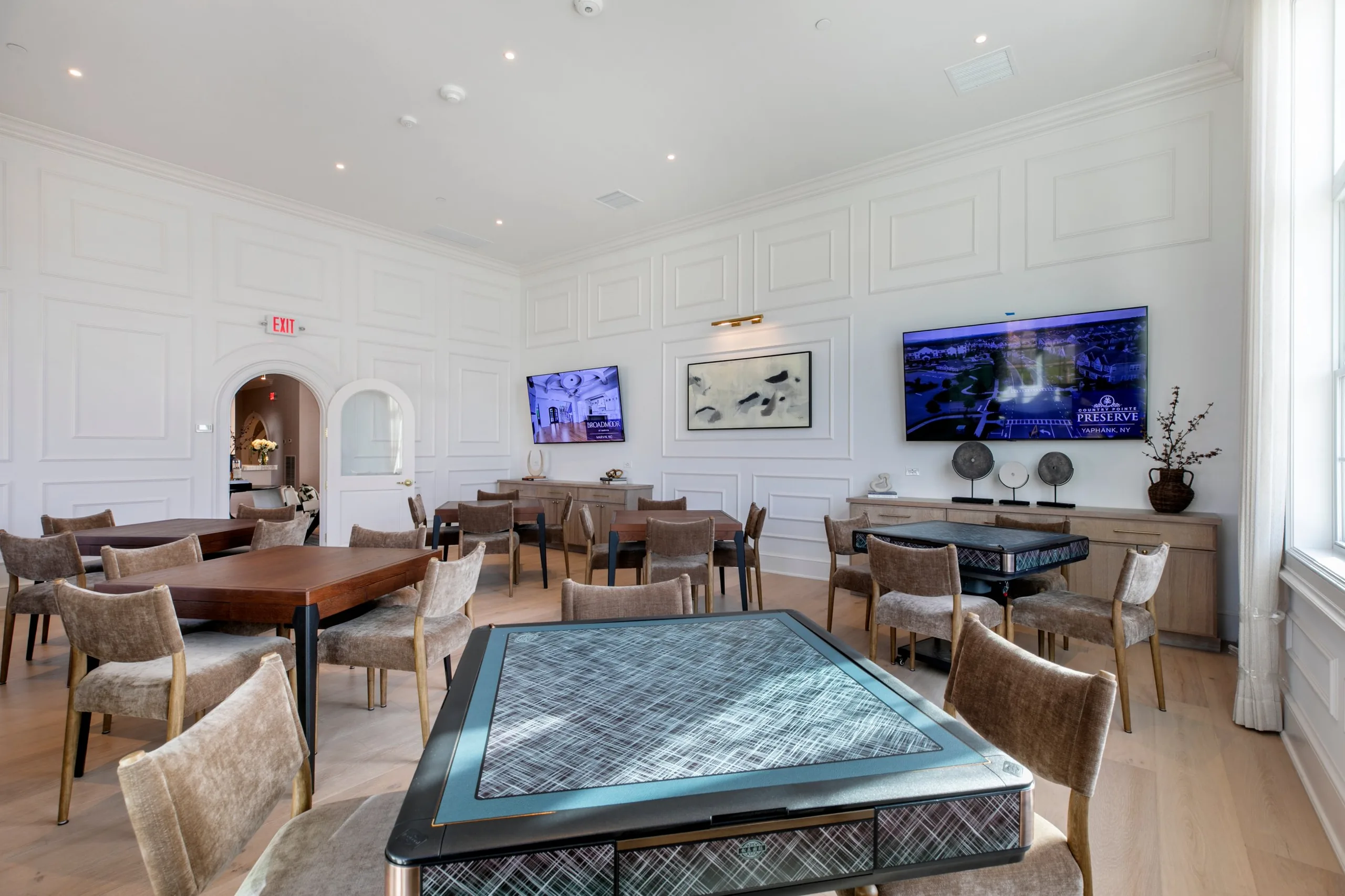 Bright card and game room with paneled white walls, wood game tables, an automatic mahjong table and upholstered chairs