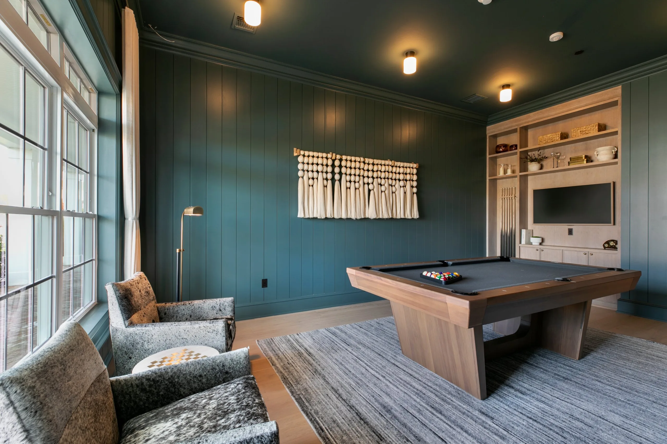 Billiards room with deep teal paneled walls, a modern wood pool table, cowhide armchairs and built-in shelving with a TV