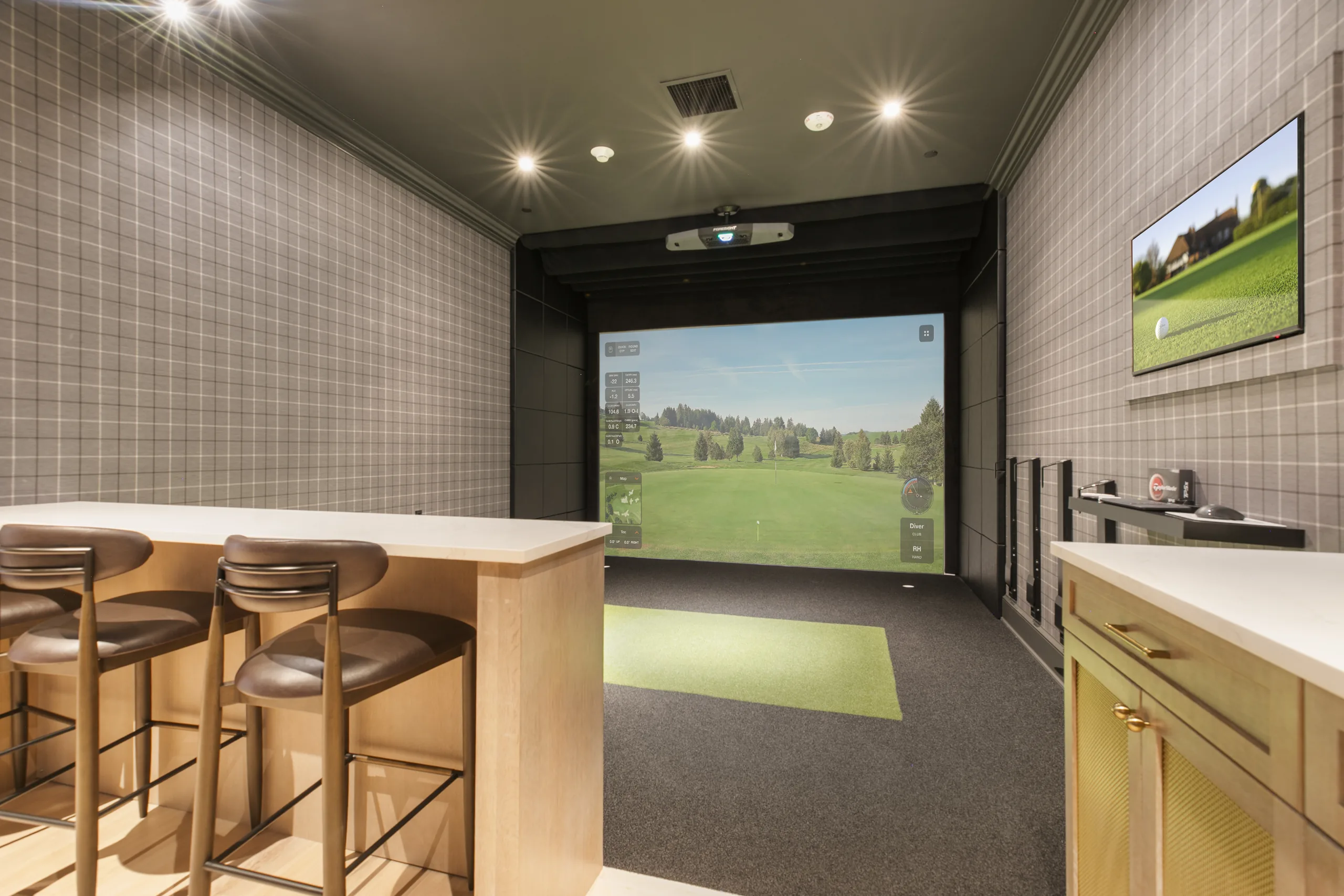 Indoor golf simulator with a full-size course projection screen, hitting mat and bar seating, in a plaid-papered room
