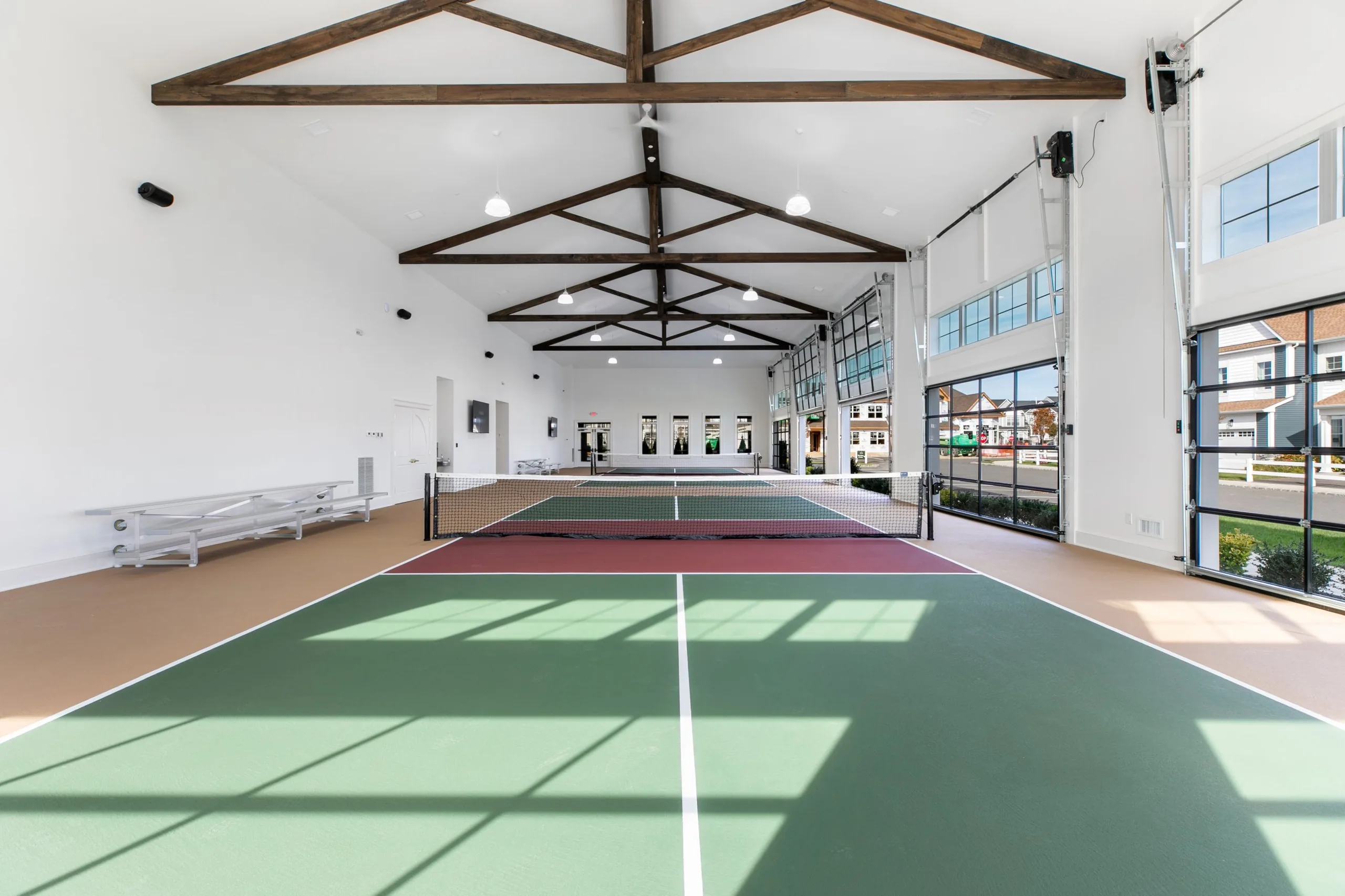 Center view of an indoor pickleball court with sunlight through glass garage doors, vaulted trusses and spectator bleachers