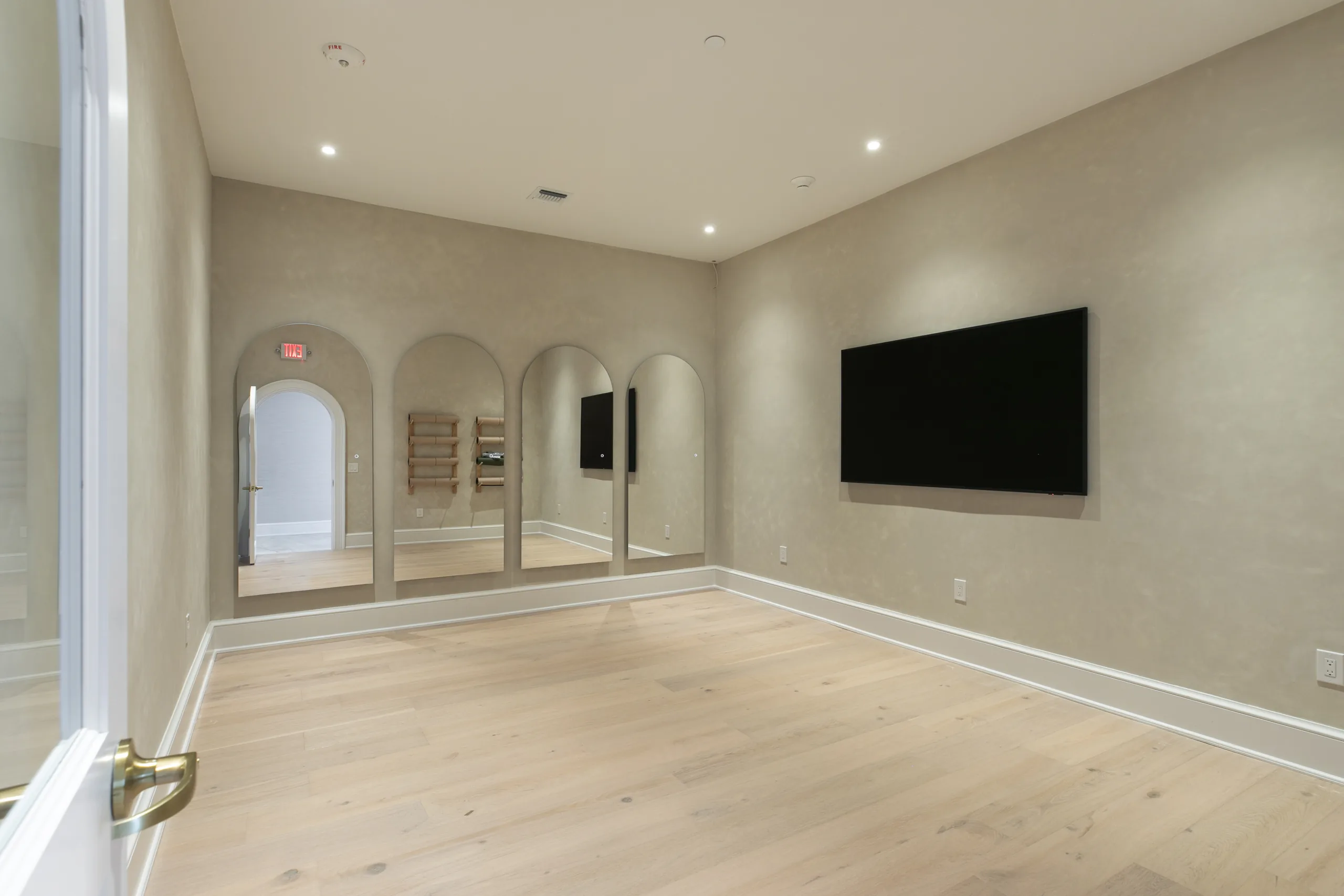 Group fitness studio with light oak floors, arched wall mirrors, a wall-mounted TV and yoga mat storage