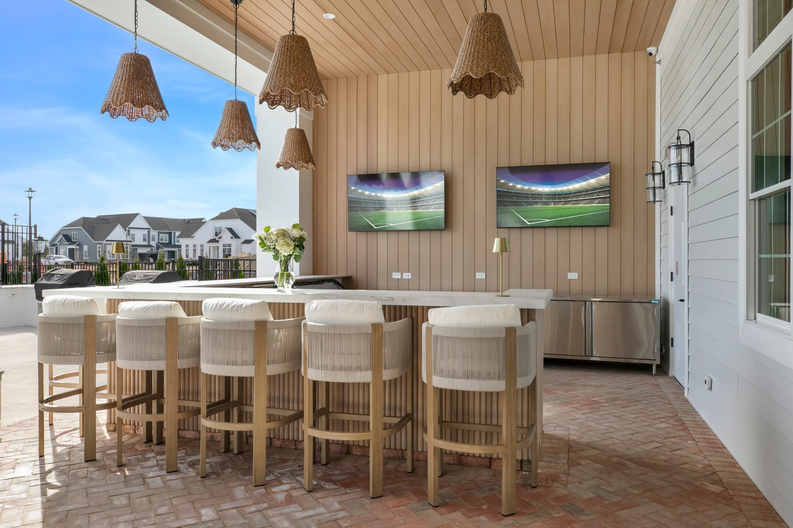 Covered outdoor bar with wood-paneled walls, scalloped woven pendants, TVs, rope-back bar stools and built-in grills
