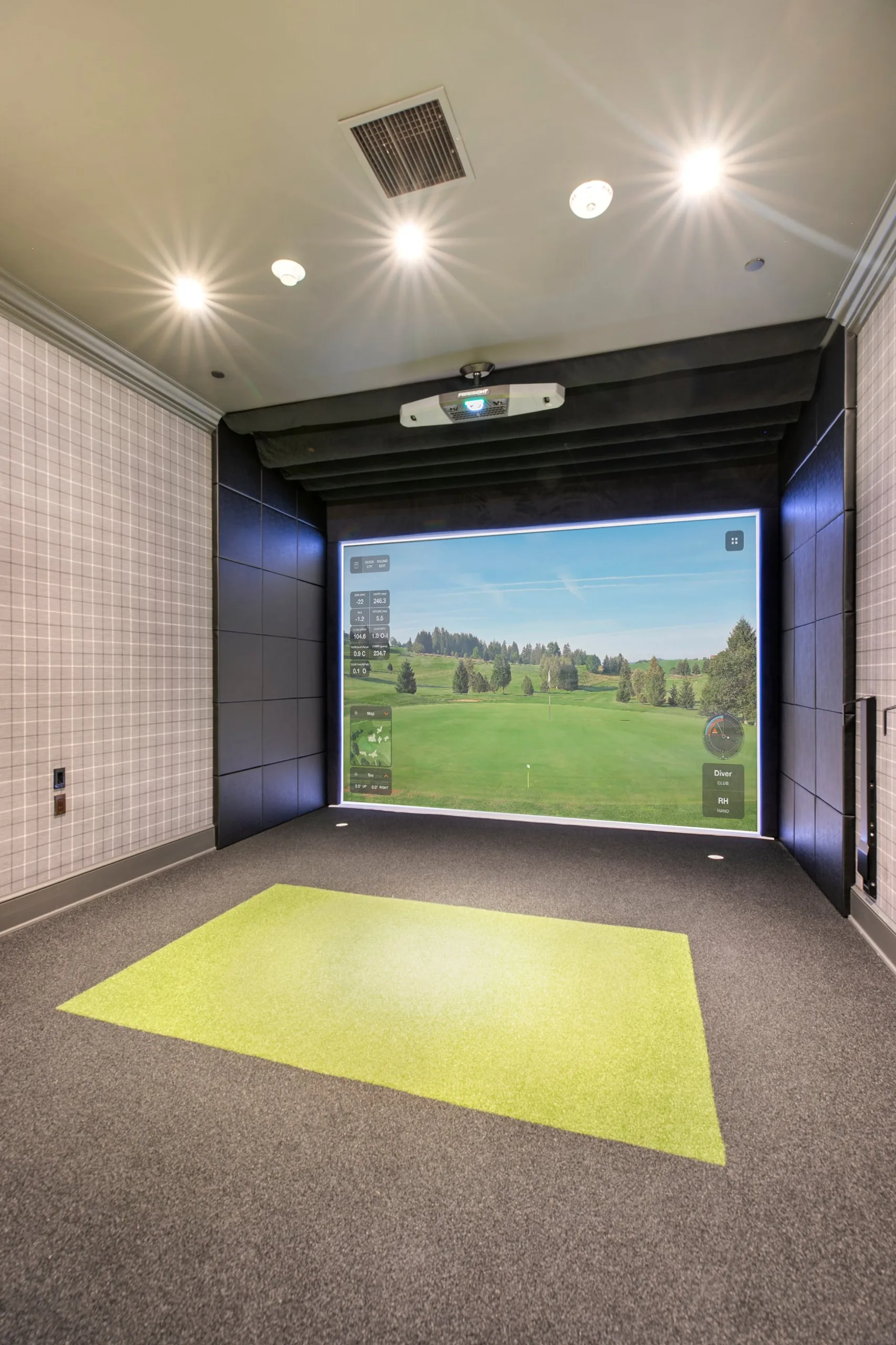 Golf simulator bay with a large course projection screen, padded side walls and a green hitting mat on charcoal carpet