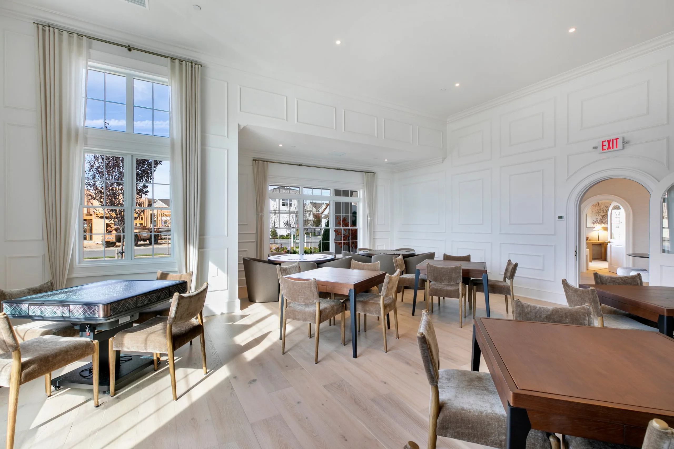 Sunlit card room with paneled white walls, tall draped windows, wood game tables, a mahjong table and upholstered chairs