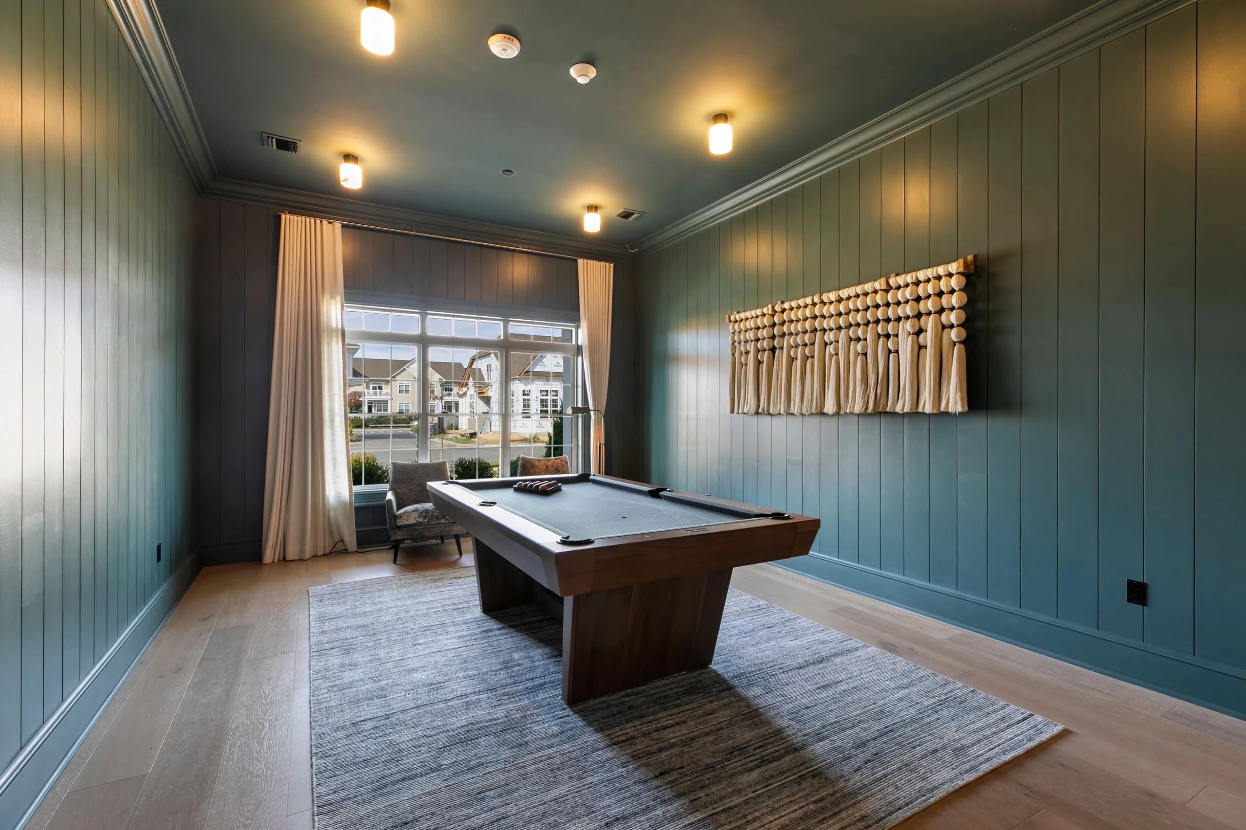 Billiards room with teal paneled walls, a modern wood pool table, a beaded tassel wall hanging and a large draped window