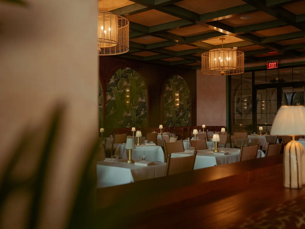 The Comet Club dining room at Vanderbilt, with rattan pendant lights, palm-leaf wall murals, white tablecloths and glowing table lamps
