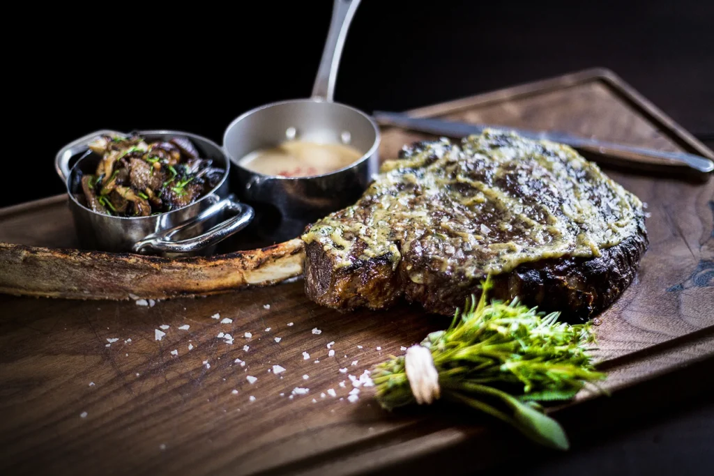 Bone-in tomahawk ribeye topped with herb butter and flaky sea salt on a wooden carving board, served with sautéed mushrooms and a side of sauce.
