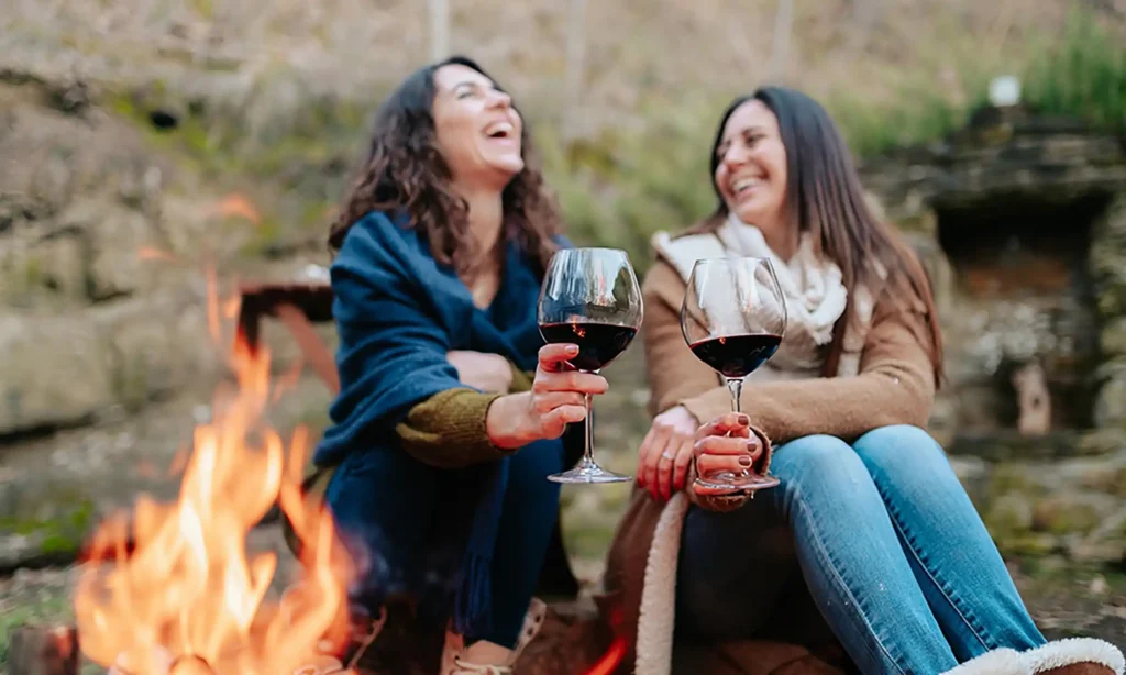 Image of 2 people enjoying a cup of wine together by a fire.